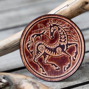 May include: A round wooden coaster with a dark brown stain features a detailed black and white illustration of a unicorn with intricate patterns. The unicorn is facing right and has a long horn.