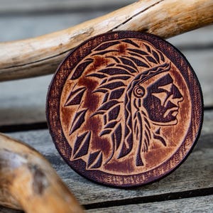 Puede incluir: Un posavasos redondo de cuero marrón con una talla detallada de un jefe nativo americano con un tocado. La talla es de color marrón oscuro, y el fondo es de un marrón más claro.