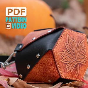 Könnte beinhalten: Eine sechseckige Ledertasche mit einer schwarzen Lederklappe und einem geprägten Ahornblatt-Design. Die Tasche hat einen Riemen und ist aus braunem und schwarzem Leder gefertigt.