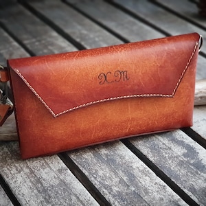 May include: A handcrafted leather clutch in shades of brown and orange, with white stitching and a wrist strap. The initials "X.M" are embossed on the front. The clutch is on a wooden surface.