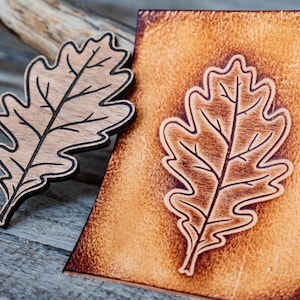 May include: A wooden oak leaf cutout with a black outline and etched details, next to a leather piece with a similar embossed leaf design. The leaf is light brown with dark brown veins. The leather is a warm, reddish-brown color.