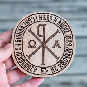 May include: A round wooden emblem with engraved Cyrillic text and a central Chi Rho symbol. The emblem is light brown and held in a hand, showcasing the detailed craftsmanship and religious theme.