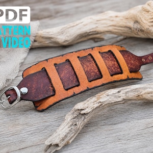 May include: A brown leather cuff bracelet featuring a layered design with a lighter tan base and darker, textured rectangular cutouts. The bracelet includes a silver-toned metal buckle closure and a pointed strap end with two small metal rivets.