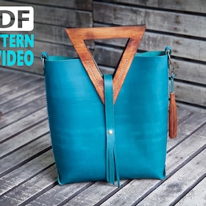 Könnte beinhalten: Eine tealfarbene Ledertasche mit einem dreieckigen Holzgriff und einem Quastenanhänger. Die Tasche hat einen Reißverschluss und einen Schultergurt. Die Tasche liegt auf einer Holzoberfläche.