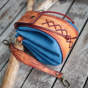 May include: A blue leather pouch with a brown leather flap and strap. The flap has a decorative stitching pattern and the strap has an intricate floral design.