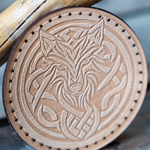 Peut inclure: Un dessous de verre rond en cuir brun clair avec un motif sculpté détaillé d'une tête de loup entourée d'un motif de nœud celtique. Le dessous de verre a une bordure cousue noire.