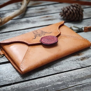 May include: A tan leather envelope-style crossbody bag with a red wax seal and a decorative design. The bag has a strap and is sitting on a wooden surface.