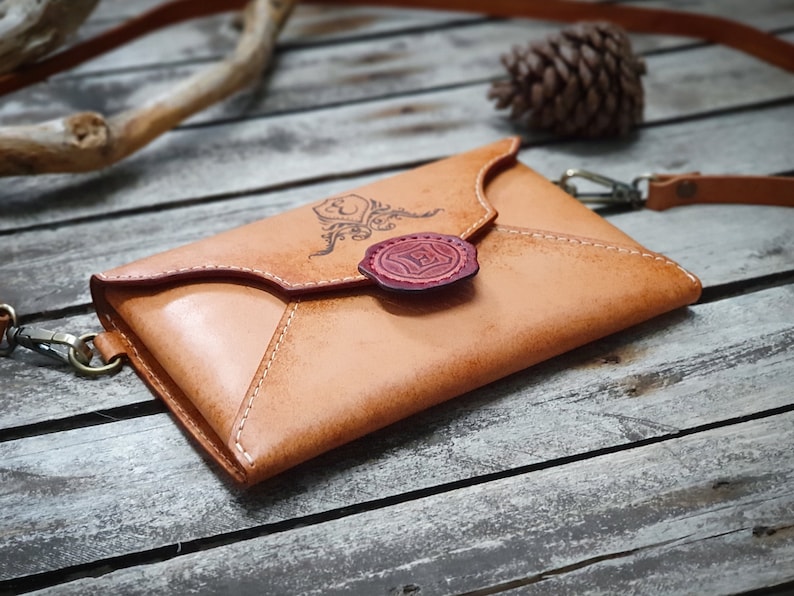 May include: A tan leather envelope-style crossbody bag with a red wax seal and a decorative design. The bag has a strap and is sitting on a wooden surface.