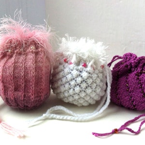 Op de afbeelding: Drie gebreide trekkoordtassen in roze, wit en paars. De roze tas heeft een pluche roze pompon bovenop. De witte tas heeft een witte pompon bovenop. De paarse tas heeft een kralenbies.