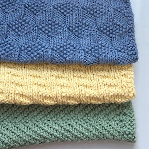 Könnte beinhalten: Drei gestrickte Geschirrtücher in verschiedenen Farben und Mustern. Das obere Geschirrtuch ist blau mit einem geometrischen Muster, das mittlere Geschirrtuch ist gelb mit einem gewebten Muster und das untere Geschirrtuch ist grün mit einem Fischgratmuster.