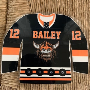 May include: Black and orange hockey jersey with the name "BAILEY" and the number "12" on the sleeves. The jersey features a Viking mascot graphic on the front. The jersey has white and orange stripes.