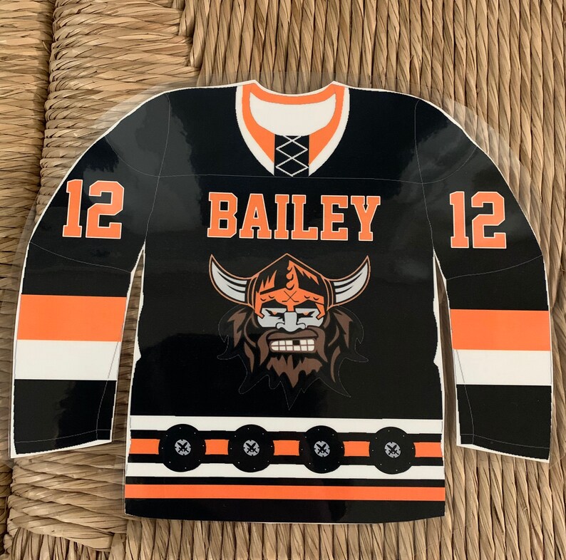 May include: Black and orange hockey jersey with the name "BAILEY" and the number "12" on the sleeves. The jersey features a Viking mascot graphic on the front. The jersey has white and orange stripes.