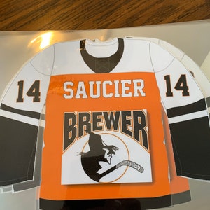May include: A hockey jersey-shaped sticker with the name "SAUCIER" and the number "14" on the front. The jersey is white, orange, and black, with a "BREWER" logo featuring a silhouette of a person playing hockey.