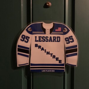 May include: A white and blue hockey jersey-shaped magnet with the name "LESSARD BRAINTREE" and the number "95" on it. The jersey has an American flag on the shoulder and the text "LAKE PLACID 2019".