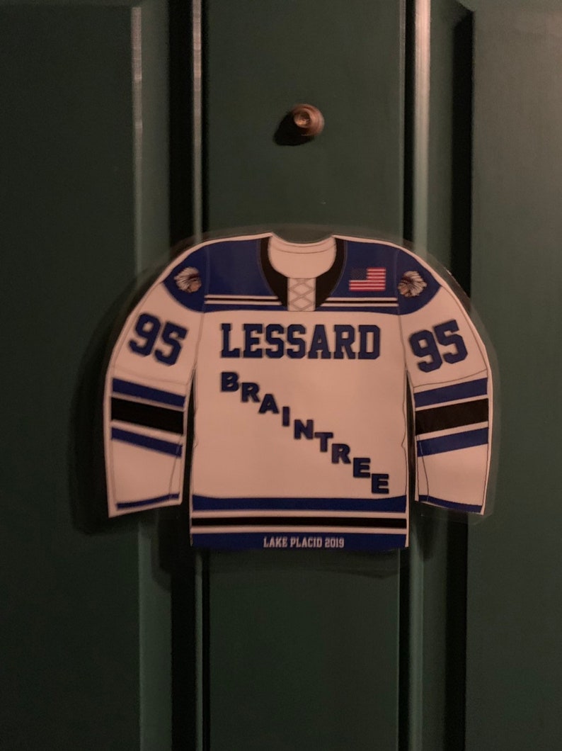 May include: A white and blue hockey jersey-shaped magnet with the name "LESSARD BRAINTREE" and the number "95" on it. The jersey has an American flag on the shoulder and the text "LAKE PLACID 2019".