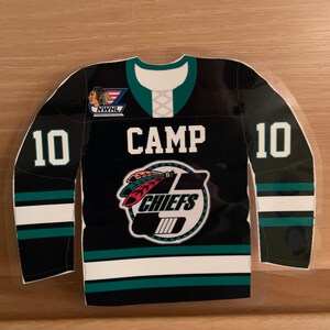 May include: A black and teal hockey jersey-shaped sticker with the word "CAMP" in white block letters. The jersey features the number "10" on both sleeves and a Chiefs logo. The NWHL logo is visible on the upper left.