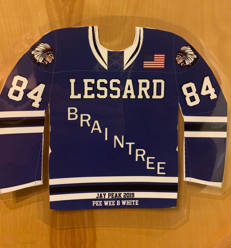 May include: A blue hockey jersey with the name "LESSARD" and "BRAINTREE" printed on it. The number "84" is on both sleeves. An American flag and a Native American head graphic are also visible. The text "JAY PEAK 2019 PEE WEE B WHITE" is printed at the bottom.