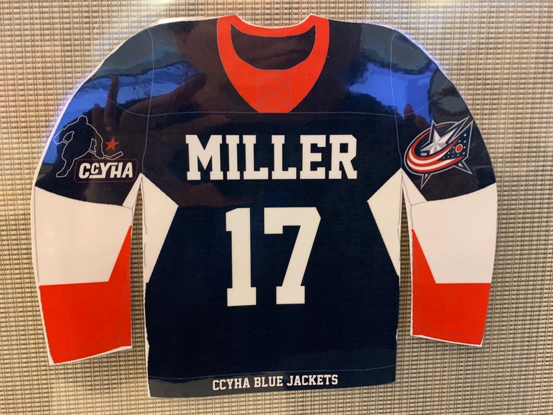 May include: A navy blue hockey jersey with white and red accents. The jersey features the name "MILLER" and the number "17" in white block letters. The team logo and "CCYHA BLUE JACKETS" are also visible.