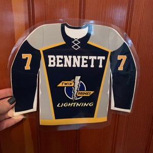 May include: A navy blue and gray hockey jersey with the name "BENNETT" and the number "7" on the back. The jersey also features the words "TWIN BRIDGES LIGHTNING" and a lightning bolt logo. The jersey is displayed against a wooden background.