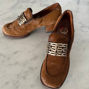 May include: A pair of brown fur loafers with a gold metal buckle detail. The loafers have a chunky heel and a rounded toe.
