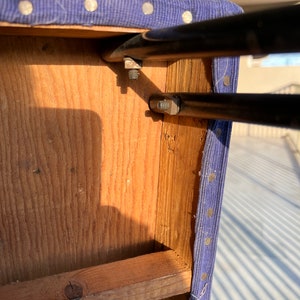 May include: Close-up of the underside of a blue upholstered stool with a wooden frame and metal supports. The metal supports are attached to the wooden frame with screws.