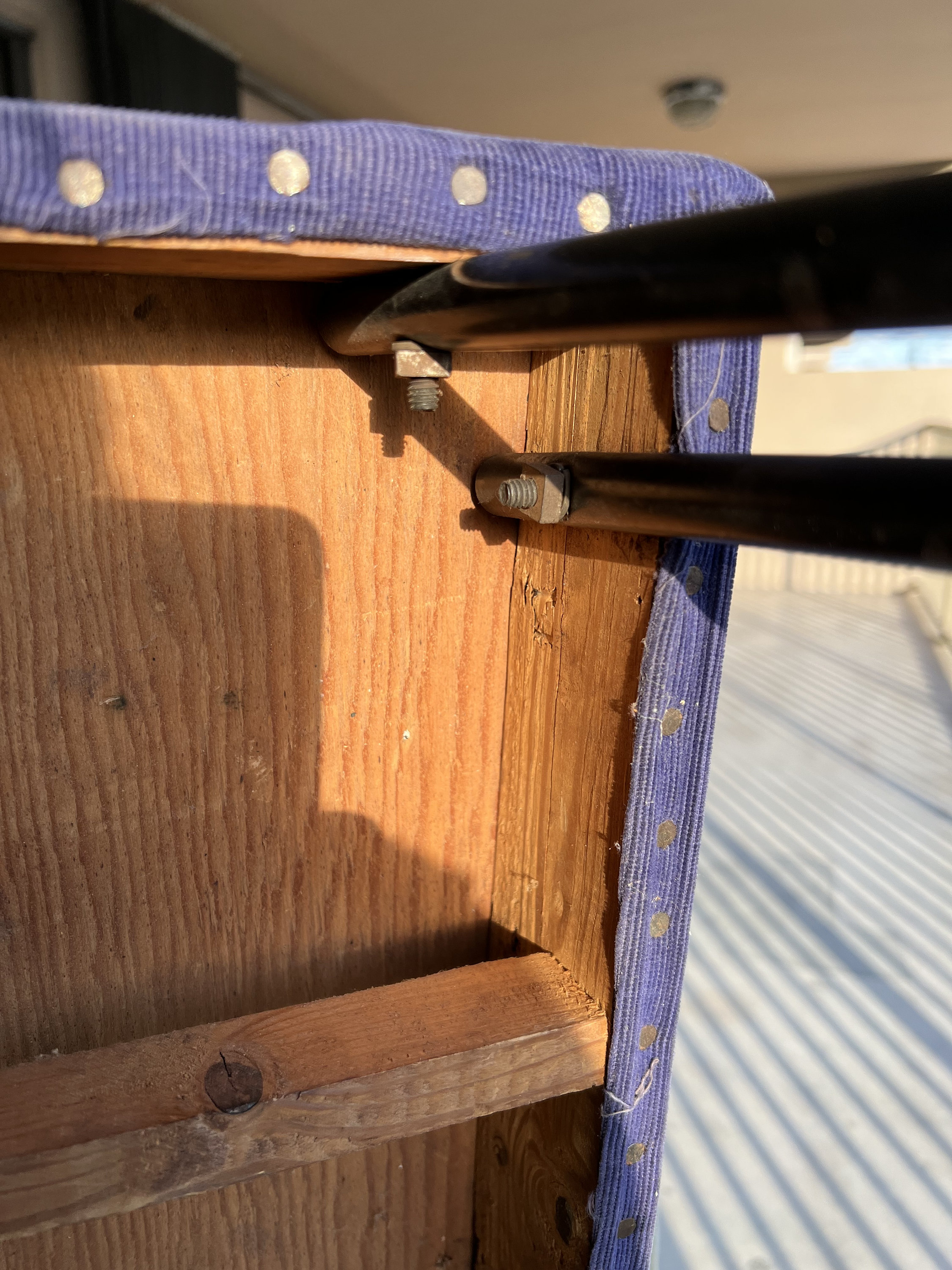 May include: Close-up of the underside of a blue upholstered stool with a wooden frame and metal supports. The metal supports are attached to the wooden frame with screws.