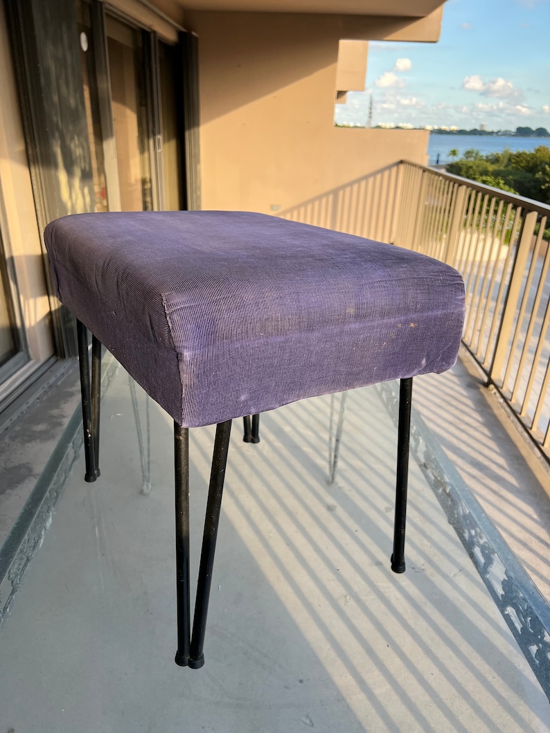 May include: A purple upholstered stool with four black metal legs. The stool is on a balcony with a view of a body of water.