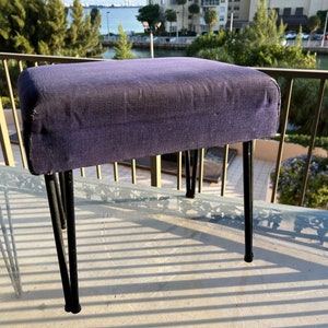 May include: A small purple upholstered footstool with black metal legs. The footstool is sitting on a glass table with a black metal frame.
