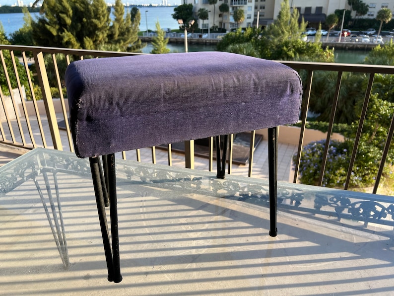 May include: A small purple upholstered footstool with black metal legs. The footstool is sitting on a glass table with a black metal frame.