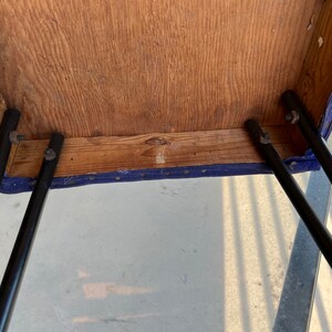 May include: The underside of a blue upholstered stool with black metal legs. The legs are attached to the stool with screws. The stool is made of wood.