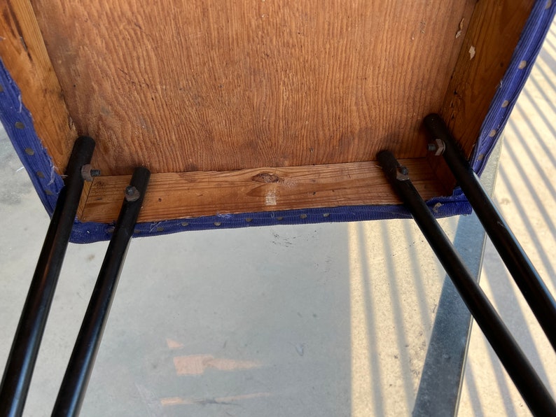 May include: The underside of a blue upholstered stool with black metal legs. The legs are attached to the stool with screws. The stool is made of wood.