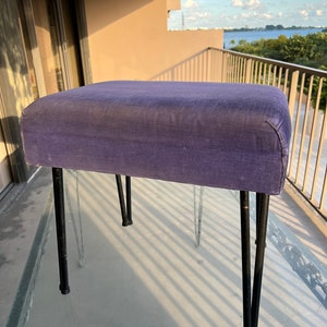 May include: A purple upholstered footstool with black metal legs. The footstool is sitting on a glass surface.