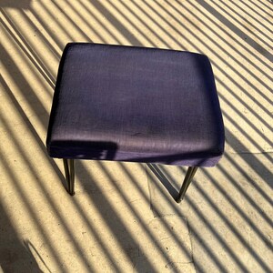 May include: A dark blue upholstered square ottoman with black metal legs. The ottoman is sitting on a concrete floor with shadows from a slatted fence.
