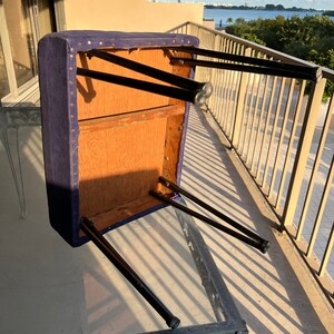 May include: A purple upholstered stool with black metal legs. The stool is flipped upside down, showing the underside of the seat and the legs.