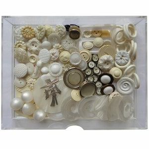 May include: A collection of white and cream coloured buttons in a clear plastic box. The buttons are of various shapes and sizes, including round, square, and oval. Some of the buttons have intricate designs, while others are plain. The buttons are arranged in a random pattern.