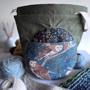May include: Olive green yarn project bag with a round owl design. The bag features a blue background with owls perched on a branch, with a floral pattern at the top. A white strap and drawstring closure are visible.