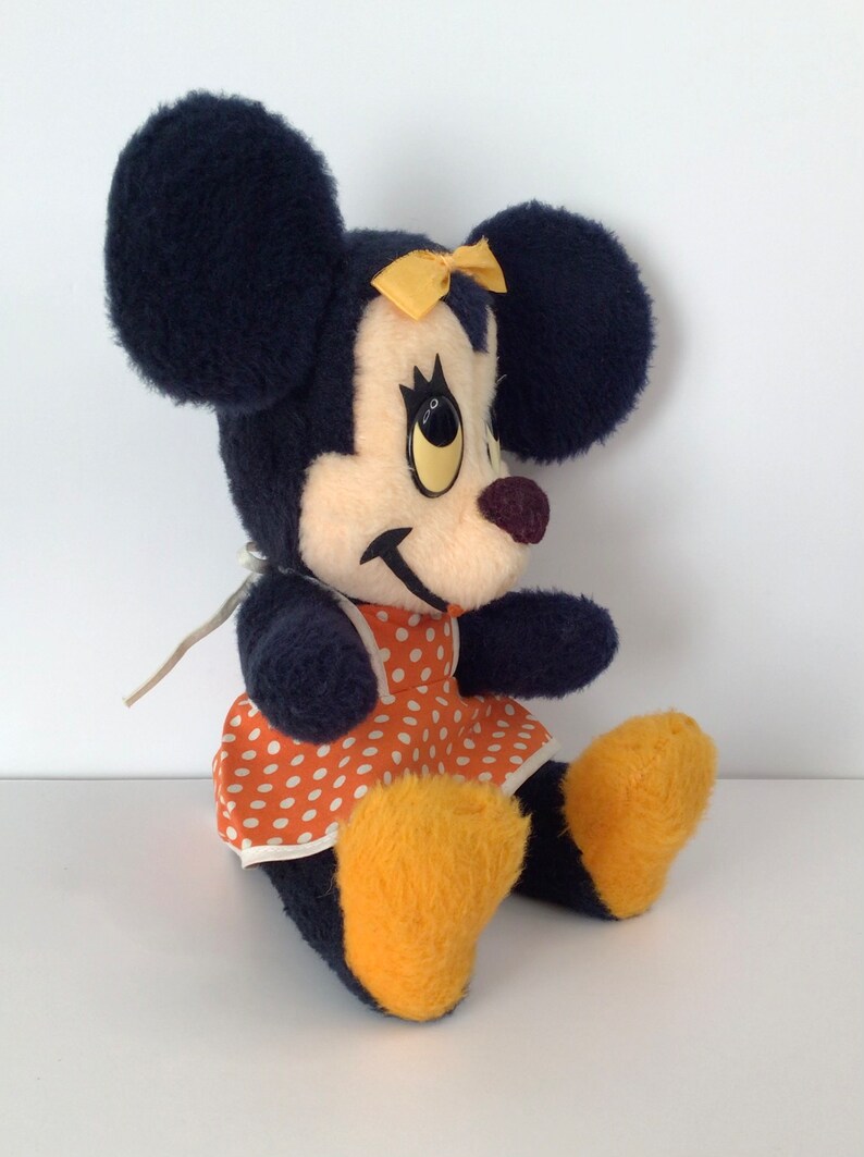 Może przedstawiać: Pluszowa zabawka przedstawiająca Minnie Mouse, dziewczynę Myszki Miki. Ma na sobie czerwoną sukienkę w białe kropki z ż&oacute;łtą kokardką. Ma duże czarne uszy i ż&oacute;łte buty.