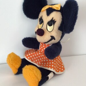 Może przedstawiać: Pluszowa zabawka przedstawiająca Minnie Mouse w granatowej sukience w białe kropki i z ż&oacute;łtą kokardką. Zabawka ma czarne uszy i ż&oacute;łte buty.