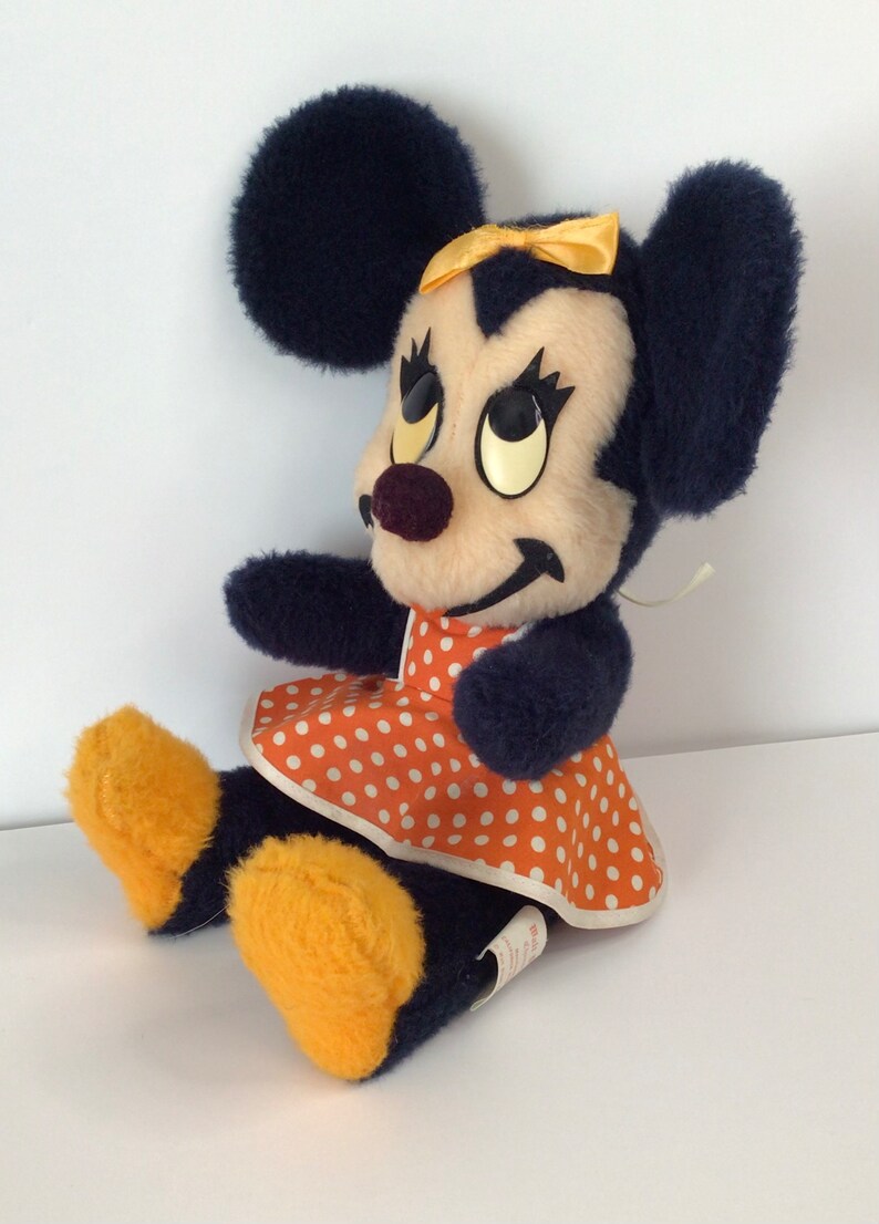 Może przedstawiać: Pluszowa zabawka przedstawiająca Minnie Mouse w granatowej sukience w białe kropki i z ż&oacute;łtą kokardką. Zabawka ma czarne uszy i ż&oacute;łte buty.