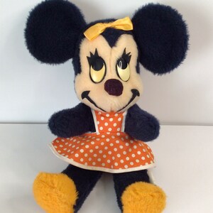Może przedstawiać: Pluszowa zabawka vintage Minnie Mouse z ż&oacute;łtą kokardką i pomarańczowo-białą sukienką w kropki. Zabawka ma lekko przerażający wyraz twarzy z dużymi, ż&oacute;łtymi oczami.