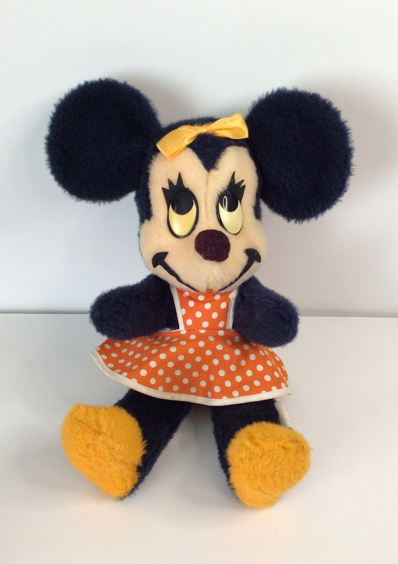 Może przedstawiać: Pluszowa zabawka vintage Minnie Mouse z ż&oacute;łtą kokardką i pomarańczowo-białą sukienką w kropki. Zabawka ma lekko przerażający wyraz twarzy z dużymi, ż&oacute;łtymi oczami.