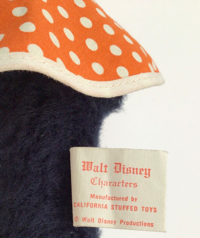 Może przedstawiać: Etykieta vintage pluszowej zabawki Walt Disney Characters z pomarańczowymi kropkami i tekstem "Walt Disney Characters, Manufactured by California Stuffed Toys, &copy; Walt Disney Productions".