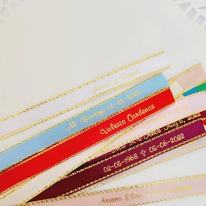 May include: A collection of colorful ribbons with gold trim and personalized text. The ribbons are white, light blue, red, pink, and burgundy. Text includes names, dates, and phrases like "Mi Bautizo" and "XV A&ntilde;os".