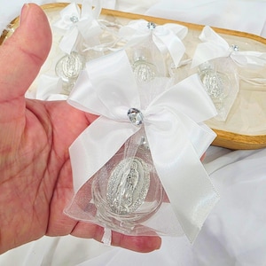 May include: Clear glass vial with a silver Virgin Mary pendant, enclosed in a sheer white pouch and tied with a white satin bow. A sparkling rhinestone accents the bow. Several similar vials are arranged in a wooden tray.