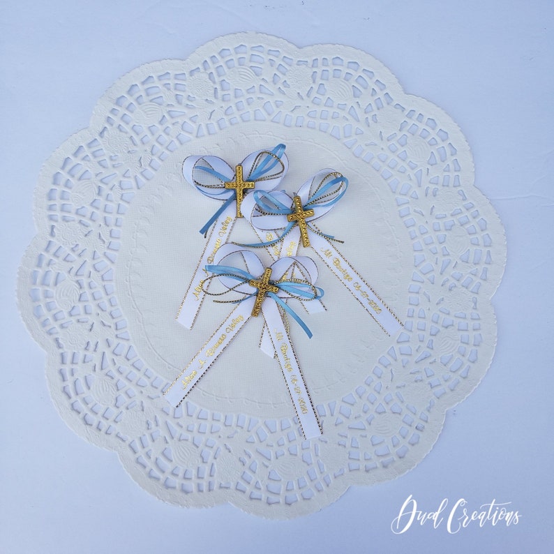 May include: Four baptism favors with white ribbons, gold crosses, and blue accents. Each favor has a ribbon with gold text. The favors are arranged on a white, lacy doily.