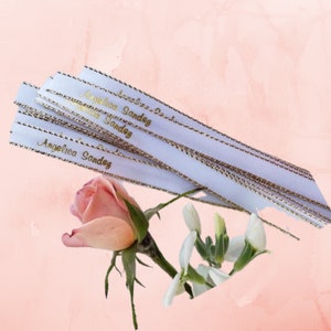 May include: White ribbons with gold text that reads "Angelica Sandez" are stacked on top of each other. The ribbons have gold trim. A pink rose and white flowers are in the foreground against a pink background.