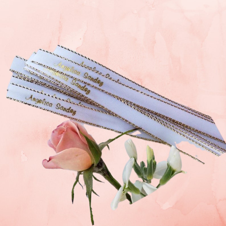 May include: White ribbons with gold text that reads "Angelica Sandez" are stacked on top of each other. The ribbons have gold trim. A pink rose and white flowers are in the foreground against a pink background.