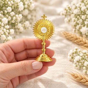 May include: A small gold-colored monstrance featuring a central white circular disc with the letters JHS inscribed in black. The object has a sunburst design with radiating rays and a small cross at the top, supported by a tiered pedestal base.