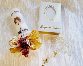 Puede incluir: Una vela blanca con un lazo dorado y un pequeño racimo de uvas rojas. La vela tiene una imagen de una niña y el texto "para Comunión" y "Andrea". Una caja blanca con el texto "Sagrada Biblia" y un rosario blanco con una cruz dorada.