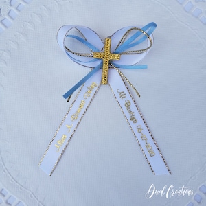 May include: A decorative bow with a gold cross centerpiece. The bow is white with light blue accents and gold trim. The ribbons have gold text that reads "Alice A. Domingo Velez" and "Mi Bautizo 06-27-2020".